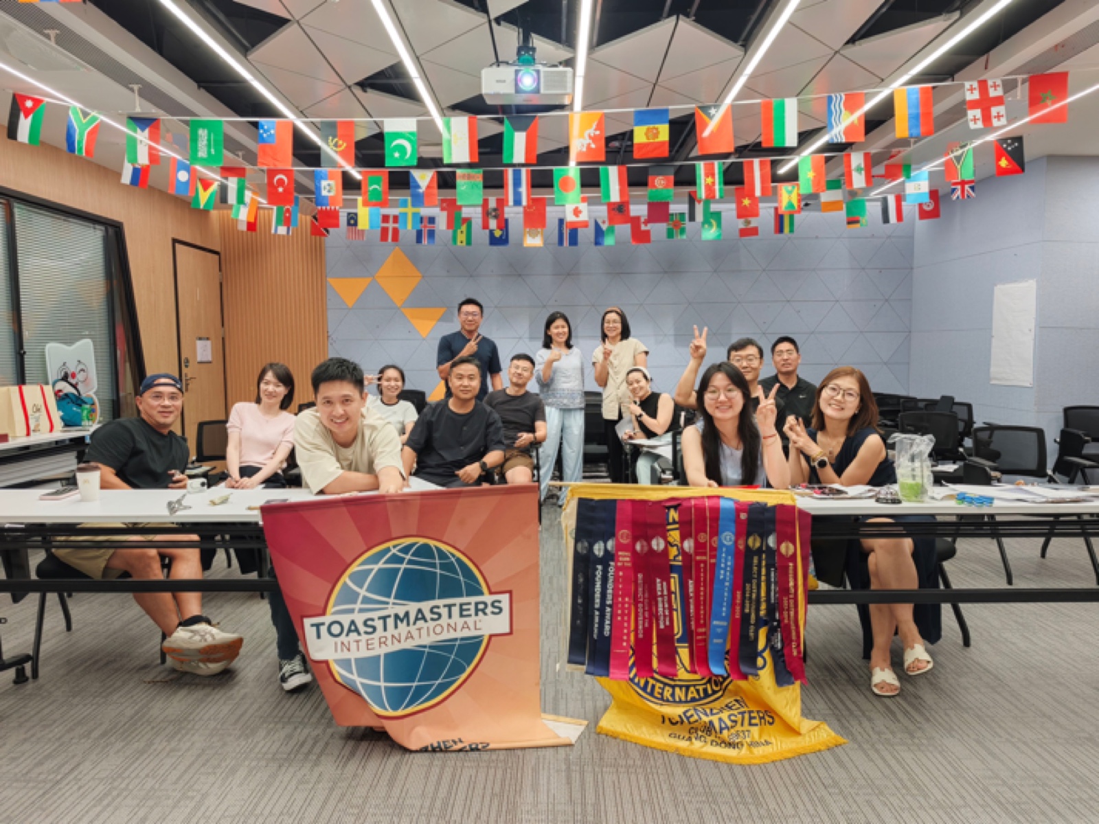 Members and guests of Shenzhen Toastmasters behind the club banner, flags of many countries strung across the ceiling